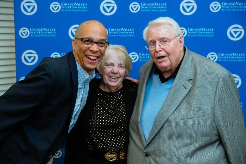 Chris Barbee and two alumni from the class of '68 pose for a photo together with the blue Grand Valley backdrop behind them at the Reunion Dinner.